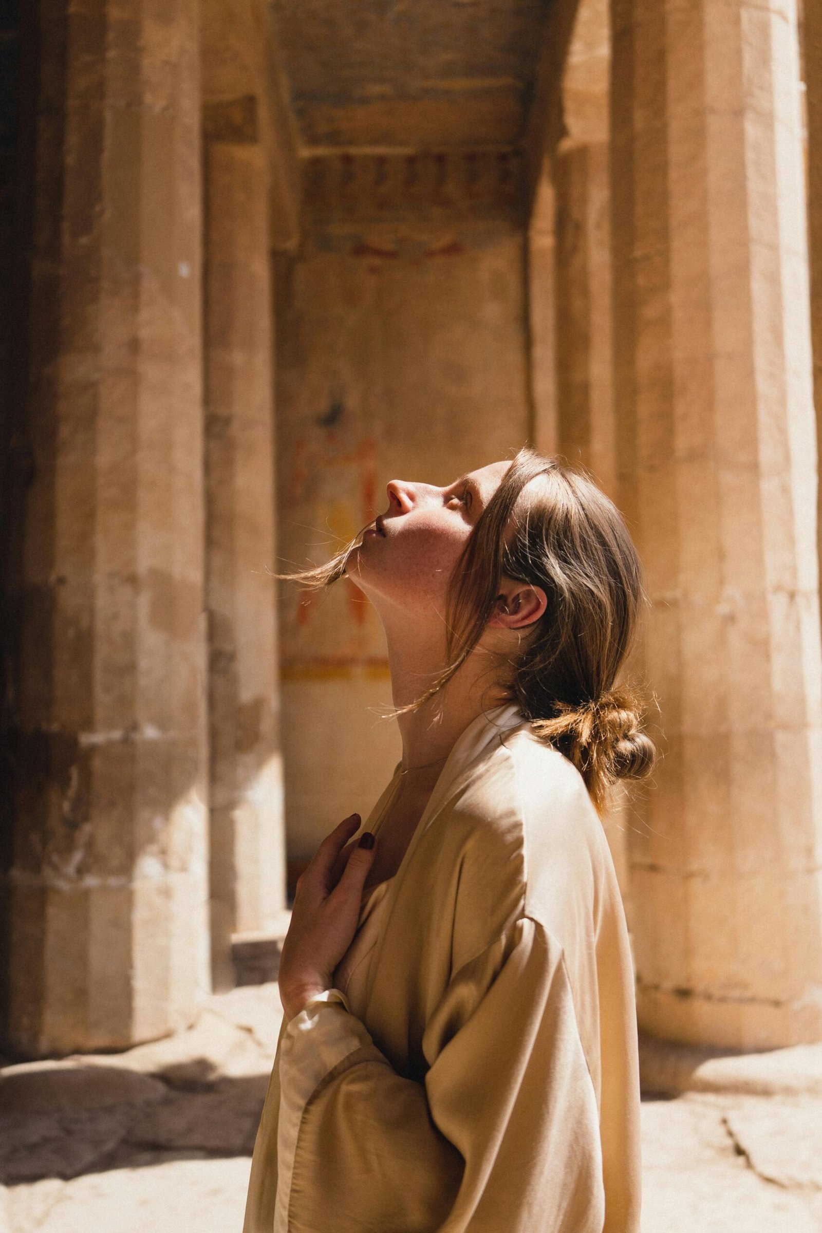 A woman in a beige coat looks up inside ancient Egyptian temple ruins.