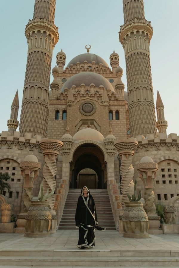 A woman in cultural attire stands before a grand mosque during the day.